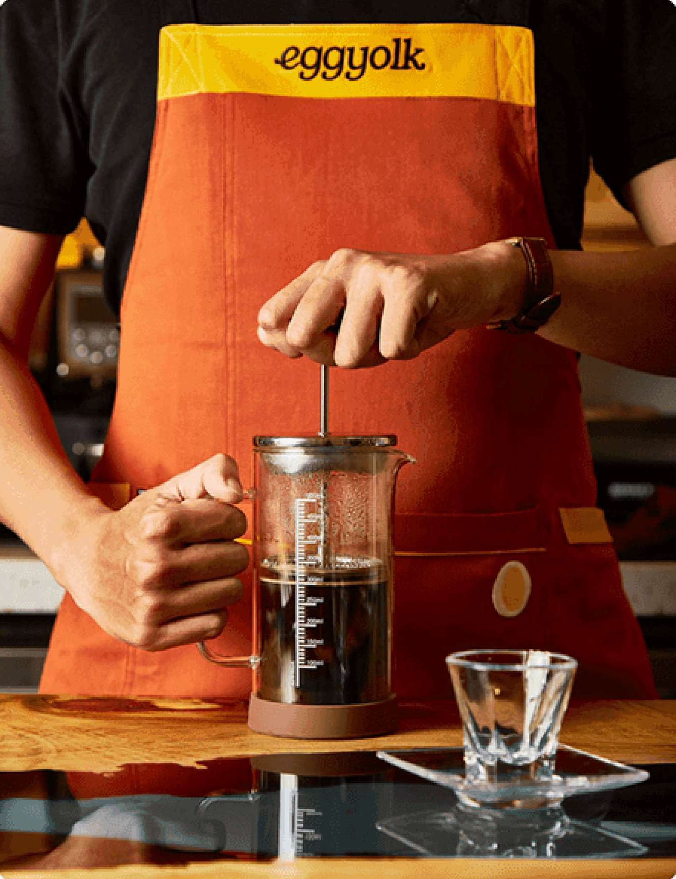 An Eggyolk barista in an orange apron brewing with a French press